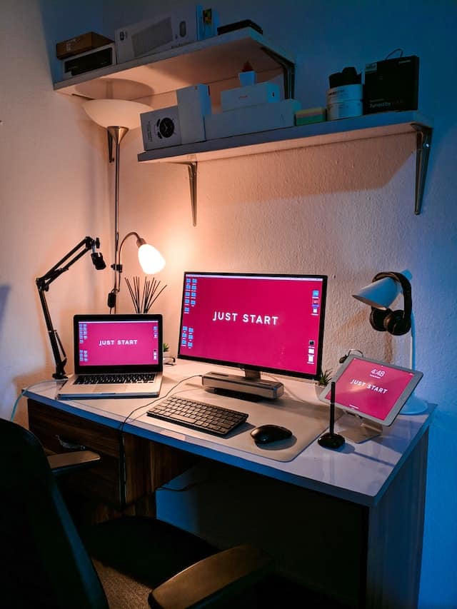 A large monitor connected to a keyboard and laptop, with an iPad also shown on a desk. They all have the words "Just Start" on the screens.