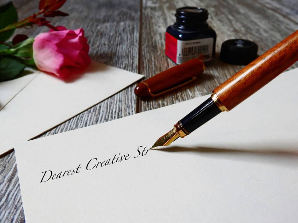 A fountain pen and writing paper surrounded by roses and ink. The pen is writing "Dearest Creative Stream,"